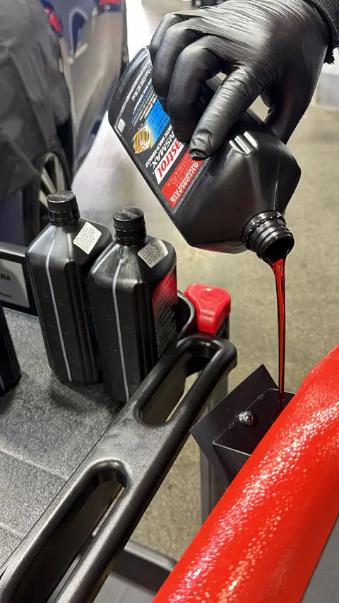 Mano con guante vierte ATF rojo de una botella Castrol Transmax, junto a otros envases sobre el carro de trabajo