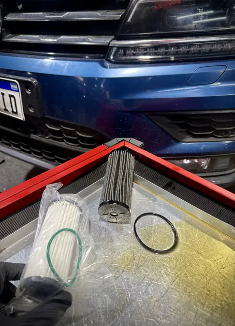 Filtro de aceite usado y filtro de reemplazo embolsado con sus juntas, sobre bandeja frente a un auto azul