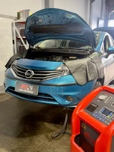 Nissan turquesa con capó abierto, cubreguardabarros y chapa F1, con la manguera del equipo en el piso