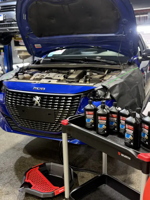 Peugeot 208 azul con capó abierto, botellas de ATF Castrol Transmax en carro Würth y bandeja de drenaje