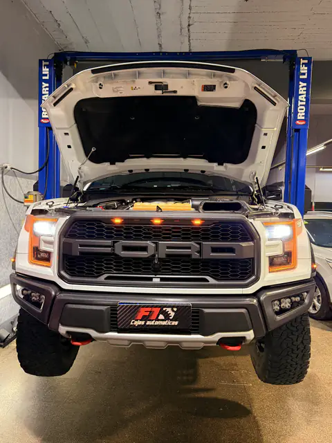 Ford Raptor blanca de frente sobre el elevador, con el capó abierto y las luces encendidas