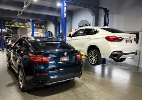 Dos BMW X6 en el taller: uno azul oscuro en el piso y otro blanco levantado en el elevador