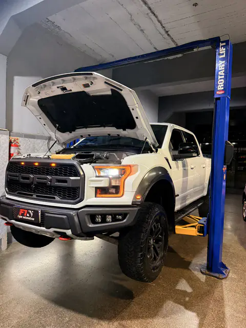 Ford Raptor blanca levantada en un elevador con el capó abierto, vista en diagonal