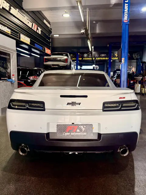 Chevrolet Camaro blanco visto desde atrás en el taller, con una pickup elevada al fondo